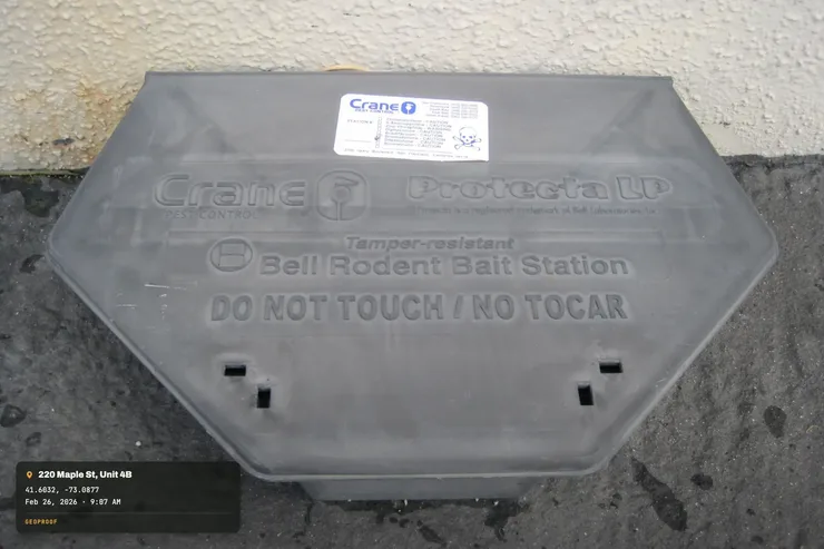 A real photo of a tamper-resistant rodent bait station against a wall base, with address and time added into the bottom-left corner as an example of GeoProof's stamp