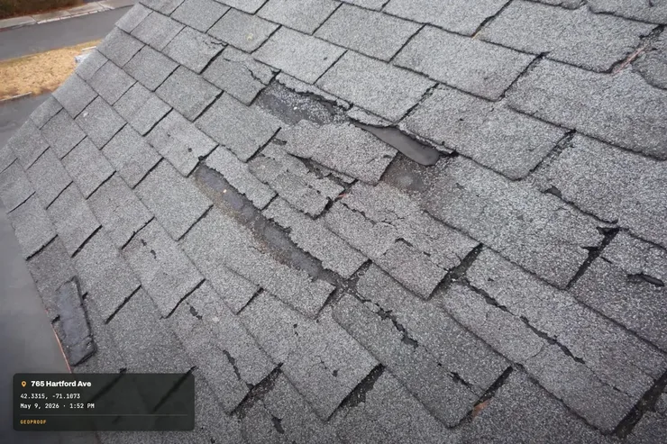 A real close-up photo of worn, damaged asphalt shingles on a roof, with GPS coordinates, address and timestamp added into the bottom-left corner as an example of GeoProof's stamp